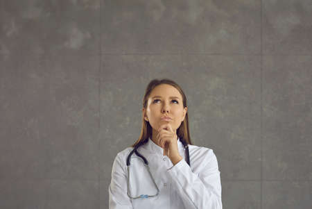 Pensive young female doctor holding her chin and looking up pondering the diagnosis. Studio shot of...