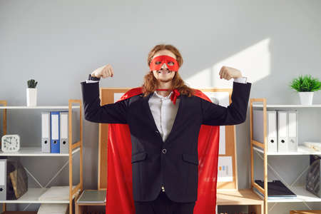 Portrait of superhero business man or employee. Funny energetic worker in red super hero mask and cape standing in office, smiling and flexing arms. Commitment to work and can-do attitude conceptの写真素材