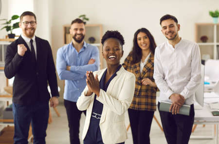 Multiethnic team celebrating success. Happy business leader or manager applauding standing in front of employees in modern office. Black woman clapping hands with multiracial colleagues in backgroundの写真素材