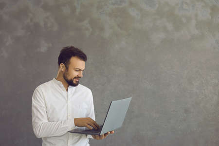 Young smiling man with a beard standing holding and typing on a laptop. An attractive man in a white...