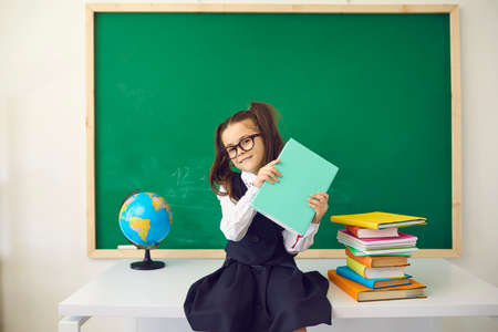 Adorable girl with ponytails sitting on teacher desk in classroom. Cute child with glasses on her...