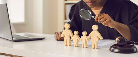 Black judge with magnifying glass looking at wooden husband, wife and children figures on desk. Court case investigation, family law, divorce, joint custody, parental rights deprivation concept.の写真素材