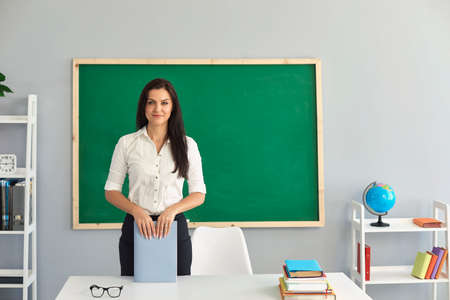 Portrait of young friendly confident smiling female teacher standing at work table in classroom against green chalkboard. Professional tutor ready for lesson. Back to school and education conceptの写真素材