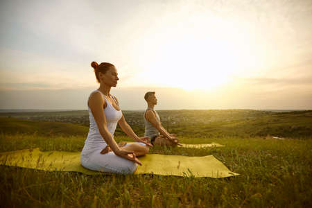 Woman and man sitting in lotus position with eyes closed, meditating and practicing yoga together on nature outdoors at sunset or sunrise on summer day. Yoga practice, healthy lifestyle conceptの写真素材