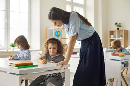 Happy teacher helping her students at the elementary school. Caring young female teacher in class tells little schoolgirl how to do schoolwork. Concept of education, teaching and knowledge.の写真素材