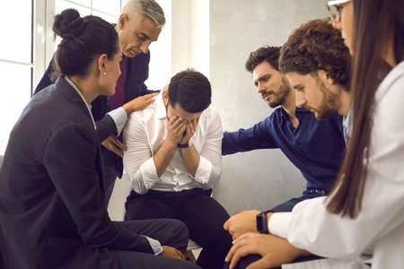 People in group therapy supporting distraught crying man, comforting him, helping him deal with grief and problems. Young man hides face and cries while telling his story sitting among other patientsの写真素材