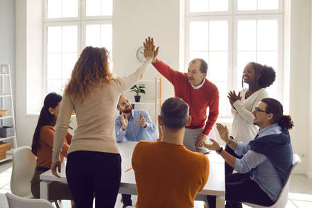 Happy young and mature coworkers and teammates high fiving each other in office meeting. Diverse business colleagues and partners celebrating successful deal. Teamwork, success and diversity conceptの写真素材
