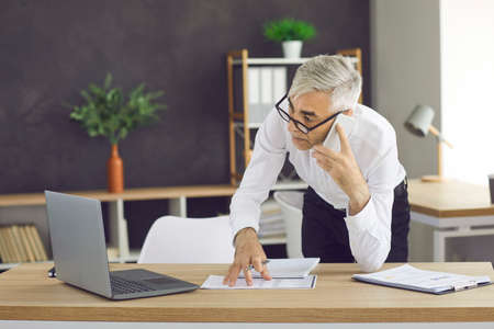 Businessman talking on a cell phone in a modern office. A man in the office looks at a laptop and talks on a cell phone. Senior man in white shirt and glasses in the office.の写真素材