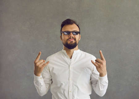 Self assured man wearing funny sunglasses. Studio shot of smirking smug looking student or businessman in white shirt and thug life glasses doing rock sign gesture standing isolated on grey backgroundの写真素材