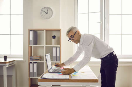 Serious business man looks at the phone in his hand and makes notes in the documents on the desk....