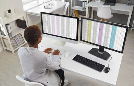 Young black woman sitting at office desk with two modern desktop computers. Financial accountant, business lady or assistant girl working with spreadsheet data lists. Back view, form above, high angleの写真素材