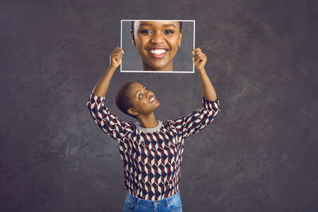 Happy pretty young black woman with short hair holding photo of her face with clear skin and perfect charming smile standing against dark grey studio background. Human emotions and dental care conceptの写真素材