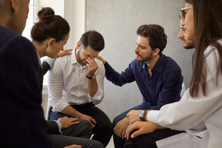Patients in group therapy meetings helping each other deal with loss and overcome grief. People comforting and supporting a depressed crying young man whos desperate after death of a close personの写真素材