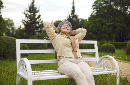 Happy delight elderly grey-haired caucasian woman looking up feeling excitement and pleasure sitting on wooden bench in city public green park outdoor in summer day. Retirement lifestyleの写真素材