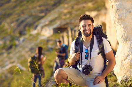 Portrait of young man with backpack and camera in mountains, copy space. Happy tourist and blurred hikers on walking tour outdoorsの写真素材