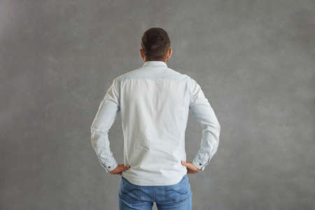 View from behind of athletic broad shouldered European man standing hands on hips against grey background. Studio backside shot of young guy with short hair in light blue denim shirt with mock up backの写真素材