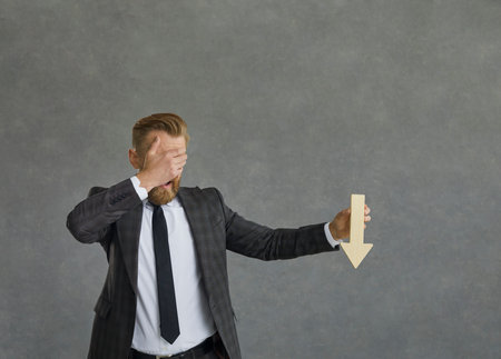 Studio shot of shocked confused businessman entrepreneur covering eyes with hand showing arrow falling down. Bankruptcy, business and financial crisis, career failure and sad fear expressionの写真素材