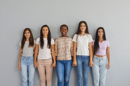 Group portrait of ordinary women of different nationalities posing against a gray wall. Smiling women in casual clothes holding hands and smiling while looking at the camera.の写真素材