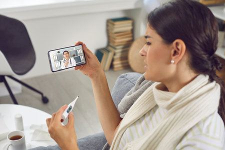 Young woman having online medical consultation via video call on mobile phone. Sick coughing lady with bad cold, flu or coronavirus fever holding thermometer and talking to telemedicine service doctorの写真素材