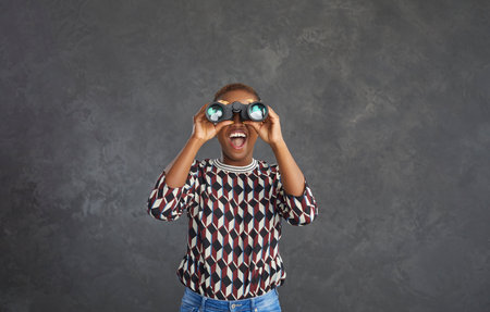 Overjoyed millennial African American woman isolated on gray studio background look in binocular glasses. Excited young biracial ethnic female watch in opera-glass. Deal, promotion, cinema concept.の写真素材