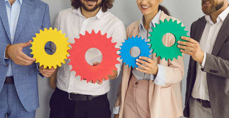 Group of happy smiling young people making chain of colorful gear wheels as symbol or metaphor for...