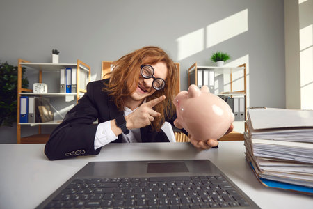 Strange crazy guy freak with round glasses talks to a piggy bank sitting at a table in front of a laptop in the office. Concept of humorous caricature on saving money and earnings.の写真素材