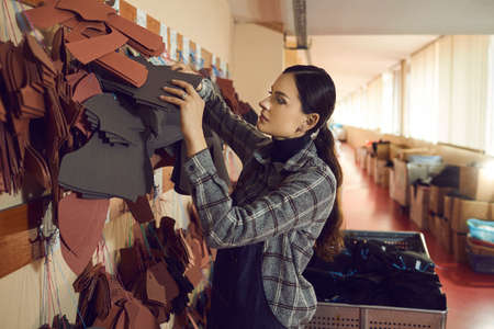 Young woman working with natural leather at shoe making factory. Female worker looking for the right footwear detail among lots of others hanging on wall peg hanger. Manufacturing industry conceptの写真素材