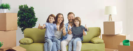 Portrait of smiling family in new home. Happy content mom, dad and kids showing thumbs up gesture sitting on sofa in room with unpacked boxes. Real estate, mortgage, buying property, ownership conceptの写真素材