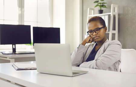 Serious woman sitting at office desk and looking at screen of modern laptop computer with thoughtful...