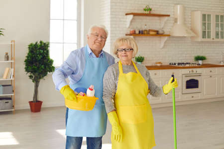Happy retired couple in aprons and gloves standing with equipment for home cleaning over spacious sunny kitchen interior. Elderly people active lifestyle, housework together conceptの写真素材