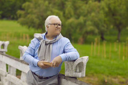Happy elderly man with a book in his hands is resting in the park outdoors. A retired man stands at the fence in the park, smiling and holding a book.の写真素材
