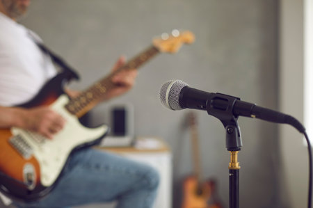 Close up of microphone in recording studio or rehearsal room, with man playing some loud music on electric or bass guitar in blurred background. Modern mic shot in soft, selective focus. Music conceptの写真素材