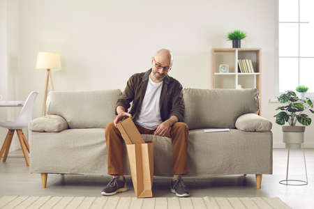 Customer takes cardboard box out of paper delivery bag. Happy bald middle aged man sitting on sofa at home and unpacking things he ordered on the Internet. Shopping online and express delivery conceptの写真素材