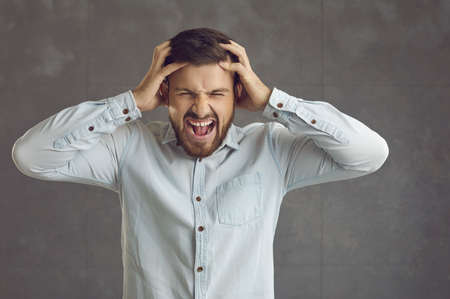 Stressed businessman going postal. Mad guy shouting and tearing hair out. Portrait of angry young man screaming and pulling hair out standing on gray studio background. Stress, rage and anger conceptの写真素材