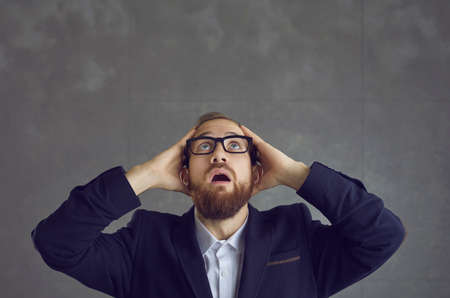 Oh no. Wow. Oh my gosh. Studio shot of scared young business man in suit and glasses looking up with mouth open and surprised face expression. Shock, fear, awe, wow effect, stunning news conceptの写真素材