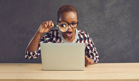 Dark-skinned woman with a surprised expression looks at the laptop screen through a magnifying...