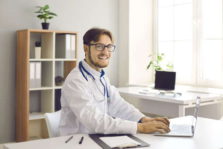 Medical workflow. Portrait of happy young male doctor working on laptop sitting at his workplace in hospital office. Medical worker who makes notes in document looks at camera and smiles sincerely.の写真素材