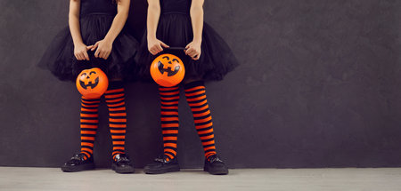 Halloween party banner. Cropped image of legs of two little girls with pumpkin candy bucket standing on black background. Children in black dresses and black and orange leggings stand near copy spaceの写真素材