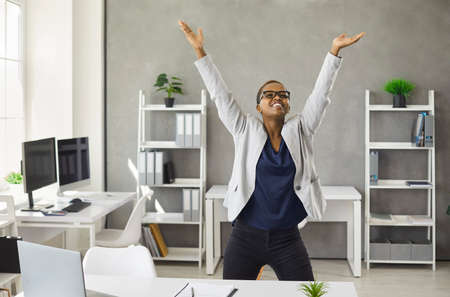 Overjoyed young African American woman businesswoman dance near desk in office celebrate job success...
