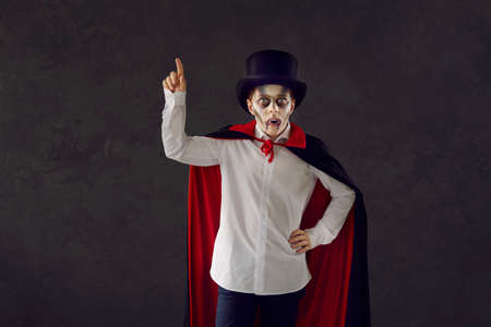 Child in vampire costume against dark studio background. Kid dressed as Dracula for Halloween party. Boy wearing top hat, red and black cape, white shirt, fake fangs and scary makeup points finger upの写真素材