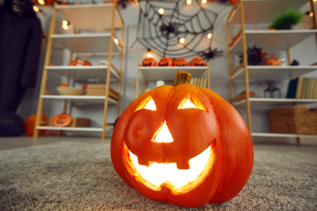 Close up of carved Jack-o-lantern standing on carpet in room with Halloween decor. Orange decorative pumpkin with carved face glows with yellow light in decorated house. Halloween party concept.の写真素材