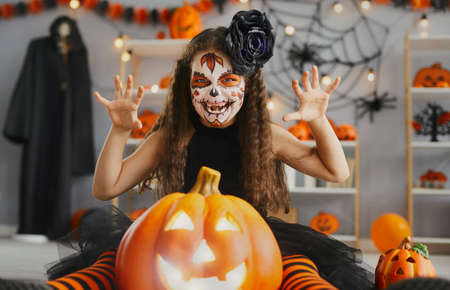Kid dressed up for Halloween party. Portrait of child in spooky costume. Little girl with beautiful skull makeup sitting on floor with Jack-o-lantern, making scary grimace and doing claw hand gestureの写真素材