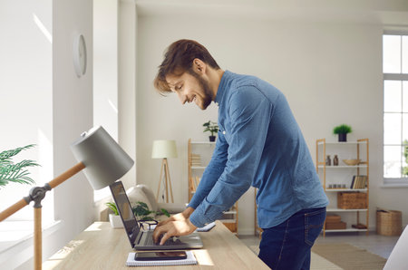 Happy young businessman or freelancer writing text on notebook while working at home office. Side view of smiling man standing at his desk searching for information on internet or writing email.の写真素材