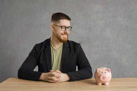 Cheerful handsome caucasian man sitting at the table and looking at the piggy bank with a smile on his face. Successful businessman sitting at the table on a gray background. Money concept.の写真素材