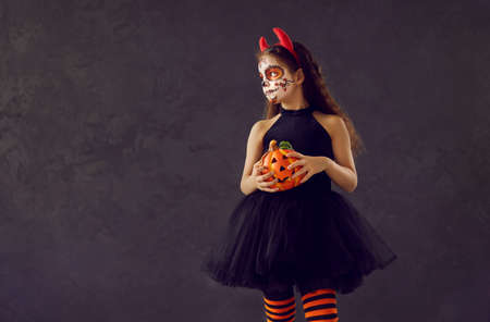 Child in Halloween costume. Portrait of little kid in scary outfit. Girl wearing beautiful black dress and skull makeup standing on dark copyspace background, holding orange pumpkin and looking awayの写真素材