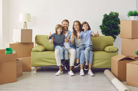 Portrait of happy family in new home. Excited content mum, dad and kids showing thumbs up gesture sitting on couch in room with unpacked boxes. Real estate, mortgage, buying house, ownership conceptの写真素材