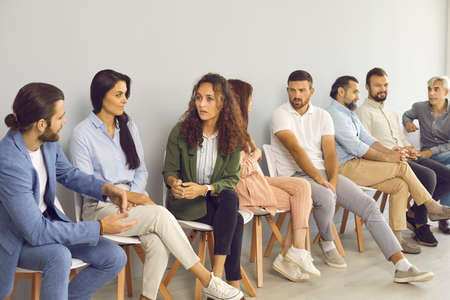 Groups of serious people of different ages talking and having discussions while sitting in queue by office wall waiting for job interview, public lecture, training session, audition or business eventの写真素材