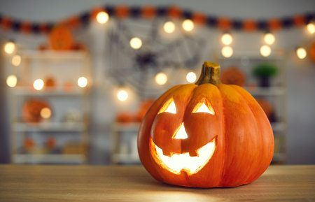 Halloween background with close up of smiley beautiful traditional carved orange Jack-O-lantern pumpkin with burning candle inside placed on wooden table in living room. Festive house decor conceptの写真素材