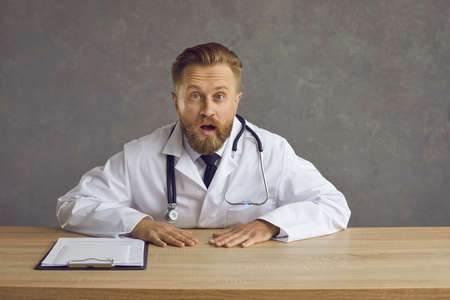 Portrait of shocked young male doctor. Man in white lab coat sitting at desk with documents and looking at camera with surprised face expressionの写真素材