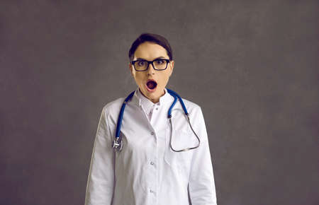 Studio shot of funny nurse or doctor surprised or scared by something. Young woman in glasses and white medical lab coat with stethoscope looking at camera with open mouth and shocked face expressionの写真素材